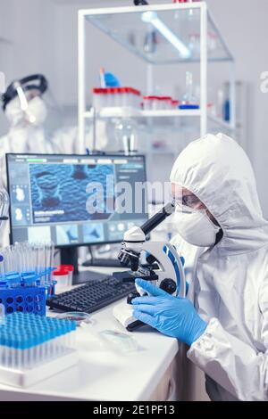 Medizinerin und Krankenschwester in PSA-Ausrüstung, die die Evolution des Coronavirus im Labor überprüft. Wissenschaftler im Schutzanzug sitzen am Arbeitsplatz mit moderner Medizintechnik während der globalen Epidemie. Stockfoto