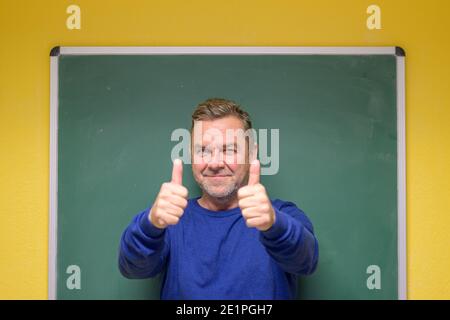 Enthusiastischer männlicher Lehrer, der mit einem strahlenden Daumen nach oben geht Lächeln Sie, als er seine Schüler vor einem gratuliert Tafel in einem Klassenzimmer Stockfoto