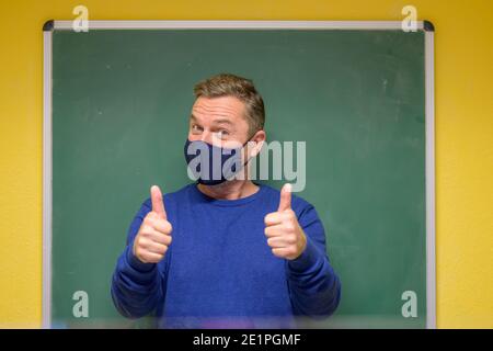 Männlicher Lehrer trägt ein Tuch Gesichtsmaske zeigt einen Erfolg Oder richtige Antwort, die einen Daumen nach oben mit beiden Händen Vor der Tafel der Klasse Stockfoto