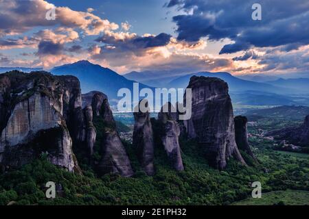 Sonnenuntergang Blick auf Meteora Tal Felsen im Vordergrund und Berge im Hintergrund. Wolken und Sonnenstrahlen fallen über das Dorf. Griechenland, UNESCO-Weltkulturerbe Stockfoto