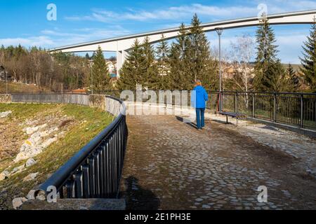 Frau in blauem Mantel, die an einem sonnigen Wintertag auf einem geländerten Damm-Damm-Damm mit Beton-Freischwinger-Viadukt als Hintergrund weggeht. Stockfoto