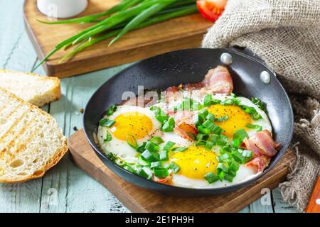 FrühlingsRühreier mit grünen Zwiebeln und Speck in einer gusseisernen Pfanne auf einem rustikalen Holzhintergrund. Stockfoto