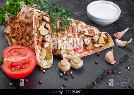 Shawarma mit Hühnerfleisch, mit Sauce, Zwiebeln, Gurken, Tomaten, Knoblauch, Kräutern und Pilzen Champignons, auf Schiefer, vor dunklem Betongrund Stockfoto