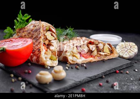 Shawarma mit Hühnerfleisch, mit Sauce, Zwiebeln, Gurken, Tomaten, Knoblauch, Kräutern und Pilzen Champignons, auf Schiefer, vor dunklem Betongrund Stockfoto