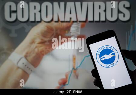 Das Logo des Brighton and Hove Albion Football Club wird angezeigt Auf einem Mobiltelefon mit einer Coronavirus-Illustration auf einem Monitor im Hintergrund Stockfoto