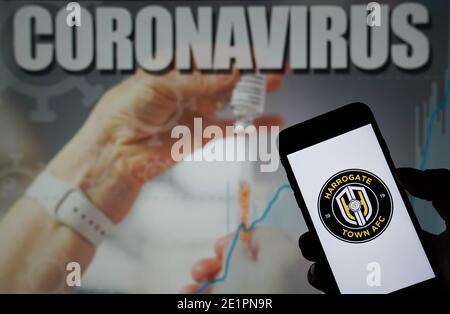 Das Harrogate Town AFC-Logo wird auf einem Handy angezeigt Telefon mit einer Coronavirus-Abbildung auf einem Monitor im Hintergrund Stockfoto