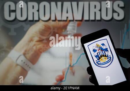 Das Huddersfield Town Football Club Logo auf einem Mobiltelefon mit Coronavirus-Illustration auf einem Monitor in Der Hintergrund Stockfoto