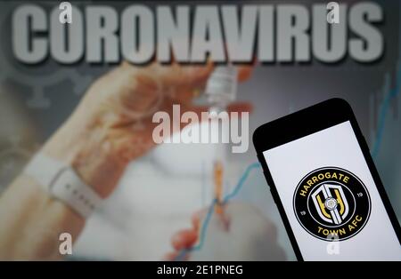 Das Harrogate Town AFC-Logo wird auf einem Handy angezeigt Telefon mit einer Coronavirus-Abbildung auf einem Monitor im Hintergrund Stockfoto