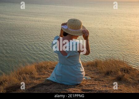 Ein Mädchen in einem Kleid und Hut sitzt auf einem Klippe am Meer Stockfoto