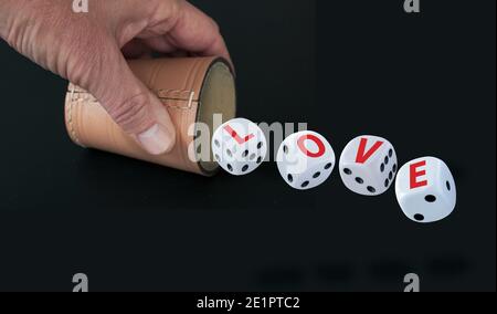 Weiße Würfel mit schwarzen Augen Zahlen Rechtschreibung LIEBE in roten Buchstaben Rollen aus einer Tombola Tasse von einer Hand isoliert auf schwarzem Hintergrund gehalten. Liebe als Stockfoto