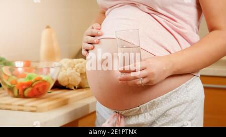 Nahaufnahme einer schwangeren Frau, die ein Glas Wasser hält und einen großen Bauch in der Küche berührt. Konzept der gesunden Lebensweise, Ernährung und Flüssigkeitszufuhr während Stockfoto