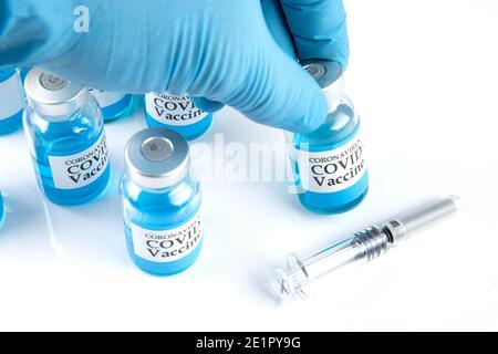 Ein Mann mit blauem Gummihandschuh, der ein Fläschchen mit blauer Flüssigkeit mit vielen weiteren Fläschchen mit Covid-19-Impfstoff und einer Glasspritze auf einem weißen Tisch hält. Stockfoto