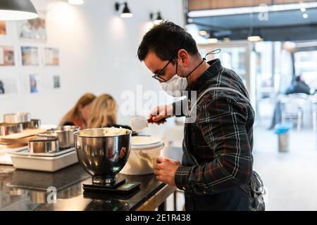 Kaukasischer Bäcker mit Maske, die einen Kuchen in einer Bäckerei zubereitet. Er setzt die Zutaten in eine Schüssel und berechnet die Mengen. Stockfoto