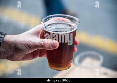 Mann, der ein kaltes Glas Bier im Freien hält. Nahaufnahme der Hand, die ein schäumendes Bier in einer Tasse hält. Hochwertige Fotos Stockfoto