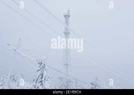 Eisbedeckter Zellenturm und elektrische Drähte über einem verschneiten Wald Auf einem Berg gegen einen Winterhimmel Stockfoto