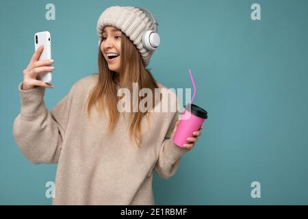 Attraktive überglücklich entzückende junge blonde Frau trägt beige Pullover und Beigefarbener Hut weiße Kopfhörer isoliert auf blauem Hintergrund halten in Hand Stockfoto