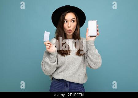 Foto von attraktiven überrascht junge Brünette Frau trägt schwarzen Hut Und grauer Pullover isoliert auf blauem Hintergrund mit Kreditkarte Und Mobiltelefon Stockfoto