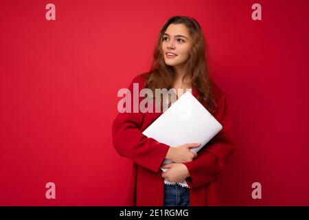 Charmante geheimnisvolle junge lockige Dame mit Netbook in roter Strickjacke Und weiße Bluse zur Seite isoliert über rot Hintergrund Stockfoto