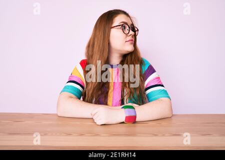 Junge Rothaarige Frau sitzt auf dem Tisch mit einer Brille, die zur Seite schaut, entspannen Profil Pose mit natürlichem Gesicht mit selbstbewussten Lächeln. Stockfoto