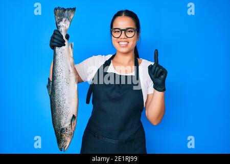 Junge schöne asiatische Mädchen Fischhändler Verkauf frische rohe Lachs lächeln mit einer Idee oder Frage Zeigefinger mit glücklichen Gesicht, Nummer eins Stockfoto