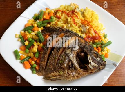 Gebratene Tilapia mit gelbem Reis, heißer Sauce und gemischtem Gemüse auf einem weißen Teller Stockfoto