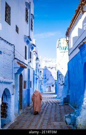 (Selektiver Fokus) EIN Mann, der die Djellaba oder Jillaba trägt, geht durch die Straßen der blauen Stadt Chefchaouen, Marokko. Stockfoto