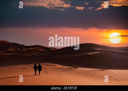 (Selektiver Fokus) Silhouette von zwei Personen, die während eines atemberaubenden Sonnenuntergangs auf den Sanddünen der Merzouga-Wüste wandern. Merzouga, Marokko. Stockfoto