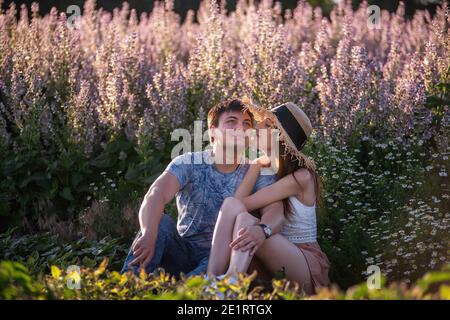 Glückliches Paar in der Liebe sitzt umarmt von einem blühenden Salbei Feld. Ein junger Mann umarmt leidenschaftlich, hält die Hände fest, blickt in die Augen eines schönen Mädchens hinein Stockfoto