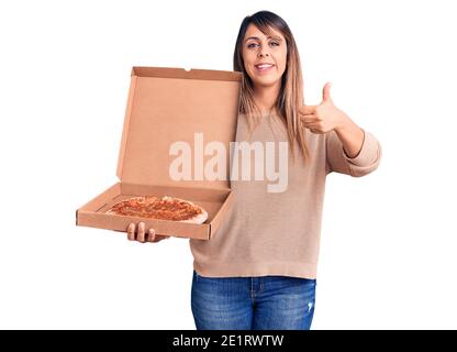 Junge schöne Frau hält Lieferung Karton mit Pizza lächelnd glücklich und positiv, Daumen nach oben tun ausgezeichnet und Genehmigung Zeichen Stockfoto