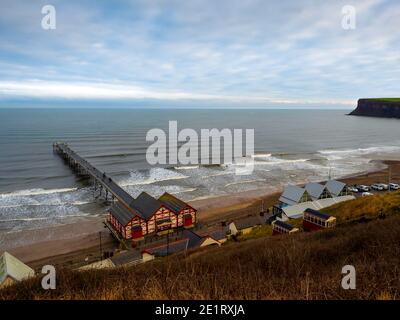 Saltburn Pier an einem Wintertag mit bewölktem Himmel Und Wetterlichtung nach einem nächtlichen Sturm und Surfern in Das Wasser Stockfoto