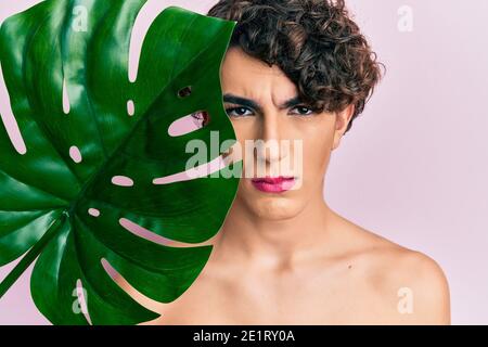 Junger Mann trägt Frau Make-up hält grüne Pflanze Blatt in der Nähe von schönen Gesicht skeptisch und nervös, runzelte wegen des Problems. Negative Pers Stockfoto