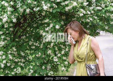 Saisonale Polynose. Frau wischt ihre Nase mit einem Taschentuch, während sie in der Nähe eines blühenden Baumes steht. Stockfoto