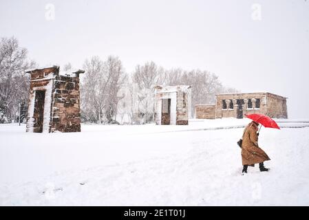 Eine Frau mit einem roten Regenschirm, die den Park Cuartel de la Montana inmitten von Schneesturm mit dem Tempel von Debod im Hintergrund überquert. Madrid, Spanien. Stockfoto