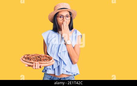 Junge schöne lateinische Mädchen trägt Sommerhut und Gläser halten Pizza über den Mund mit der Hand, schockiert und Angst vor Fehler. Überrascht Ausdruck Stockfoto