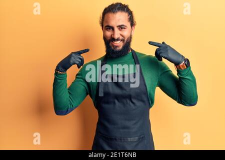 Junger arabischer Mann mit Friseurschürze, der selbstbewusst mit einem Lächeln im Gesicht aussieht und sich mit stolzen und glücklichen Fingern zeigt. Stockfoto