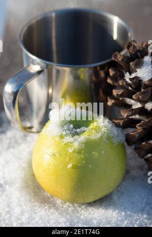 Grüner Apfel im Schnee mit einem Zedernkegel und einem Metallbecher für einen heißen Drink. Winter für einen Snack in der Natur Stockfoto