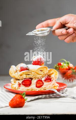 Ein Stück Erdbeerkuchen auf einem roten Teller. Hand des Mannes mit Sieb Puderzucker von oben. Grauer Hintergrund, vertikale Komposition Stockfoto