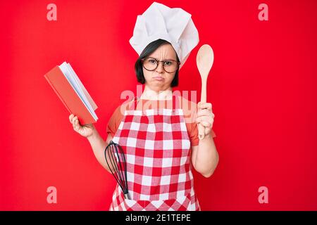 Brunette Frau mit Down-Syndrom tragen professionelle Bäcker Schürze Lesen Kochen Rezept Buch deprimiert und sorgen für Not, Weinen wütend und afra Stockfoto