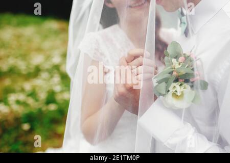 Bräutigam in weißem Hemd mit Fliege und Boutonniere hält Braut Hand unter Schleier Stockfoto