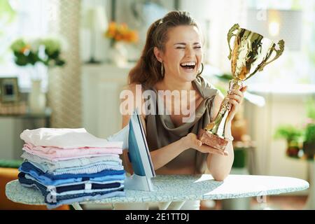 Lächelnd stilvolle Mittelalterhausfrau in Seidenbluse und beige Hosen mit Dampfbügeleisen, Bügelbrett, Stapel gefalteter gebügelter Kleidung und Gold-Kelch in m Stockfoto