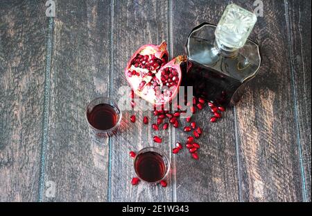 Dunkelroter Likör wird aus einer Flasche in zwei Gläser auf einem Holztisch gegossen, neben reifem Granatapfel und seinen Samen. Flach liegend. Stockfoto