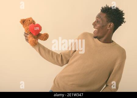 Profil anzeigen von Jungen glücklich schwarzen afrikanischen Mann lächelnd, während bei Teddybär mit Herz und Liebe Zeichen gegen weißen Hintergrund suchen Stockfoto