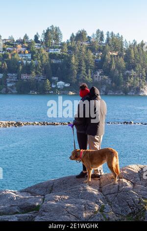 Rückansicht des jungen Mannes und der Frau steht auf einer felsigen Klippe, die sich und ihren Hund neben sich umarmt. Kalter Herbsttag. Selektiver Fokus, trav Stockfoto