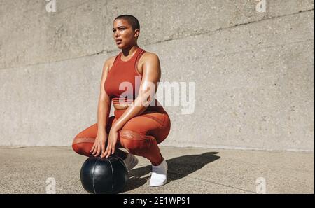 Fitness-Frau, die sich mit Medizinball auf dem Boden hockt. Damen in Übergröße in Sportbekleidung mit Medizinball, der wegblickt. Stockfoto