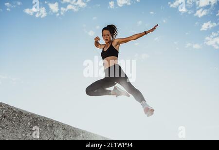 Fitness-Frau, die von der Wand springt als Trainingsroutine. Tiefwinkelansicht einer Sportlerin in Sportbekleidung, die morgens im Freien trainiert. Stockfoto