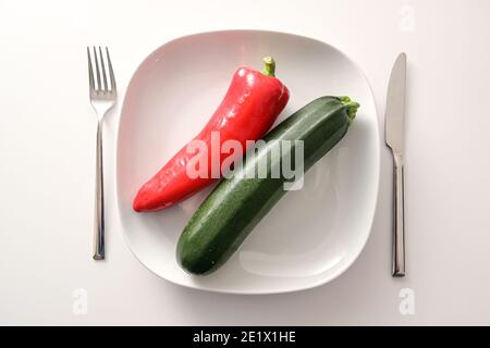 Roher roter, spitzer Pfeffer und Zucchini auf weißem Teller und Besteck auf hellem Hintergrund, gesunde Ernährung mit mediterranem Gemüse, um Gewicht zu verlieren Stockfoto