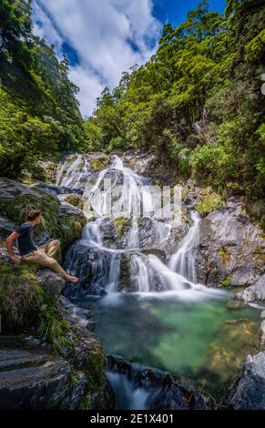 Junger Mann sitzt an einem Wasserfall, Fantail Falls, Makarora River, Wanaka, Westküste, Südinsel, Neuseeland Stockfoto