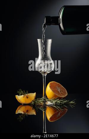 Gin wird in ein kleines Glas aus einer antiken Flasche aus dunklem Glas gegossen. Ein starkes alkoholisches Getränk mit Zitrone und Rosmarin auf einem schwarzen reflektierenden Hintergrund Stockfoto