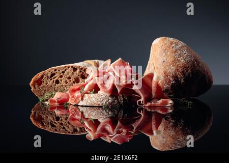 Prosciutto mit Ciabatta und Thymian auf schwarzem reflektierenden Hintergrund. Stockfoto