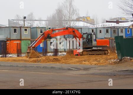 Ein großer orangefarbener Bagger auf breiten Eisenbahnen steht auf einer Baustelle auf dem Sand vor dem Hintergrund von temporären Bauerhäusern. Stockfoto